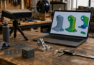 A workshop bench with a small 3D scanner, calibration target, mechanical part, and laptop mesh inspection view.