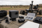 A rugged action camera field documentation kit with mounts, batteries, tools, and a small robot chassis on a neutral work surface.