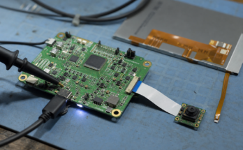 Macro electronics bench view of a generic multimedia microcontroller board with camera module, ribbon cable, and test probe