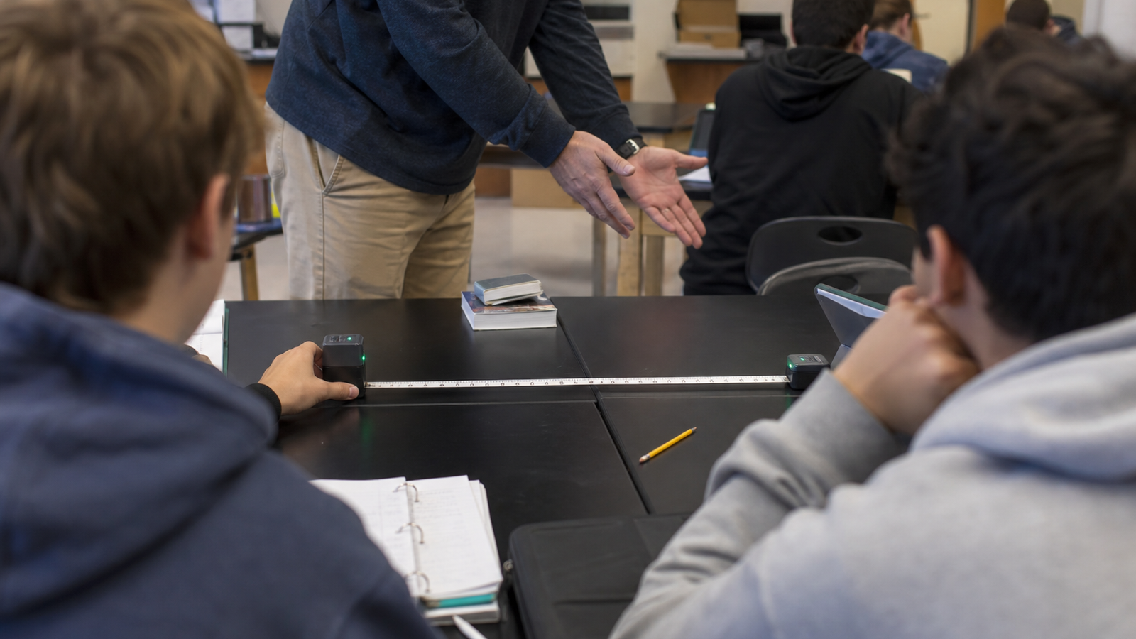 STEM classroom scene with students measuring distance between two small beacons on a table, teacher supervising, no faces pro
