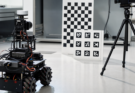  small wheeled robot facing a printed checkerboard and ArUco board under soft side lighting, tripod camera, no logos, no read