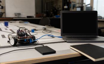Student robotics lab debugging scene: small wheeled robot on test mat, external SSD, unbranded laptop off to side with blank screen, Ethernet cable an