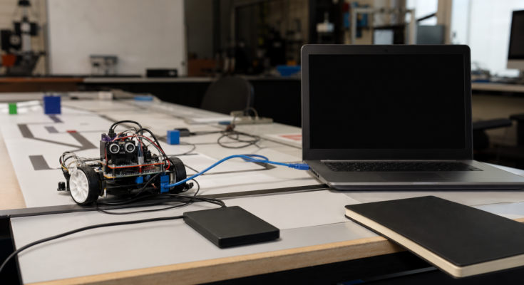Student robotics lab debugging scene: small wheeled robot on test mat, external SSD, unbranded laptop off to side with blank screen, Ethernet cable an
