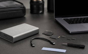 Professional field-media storage kit on a table: unbranded aluminum NVMe enclosure, short certified cable, camera cards, laptop off to far side with blank 