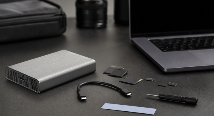 Professional field-media storage kit on a table: unbranded aluminum NVMe enclosure, short certified cable, camera cards, laptop off to far side with blank 
