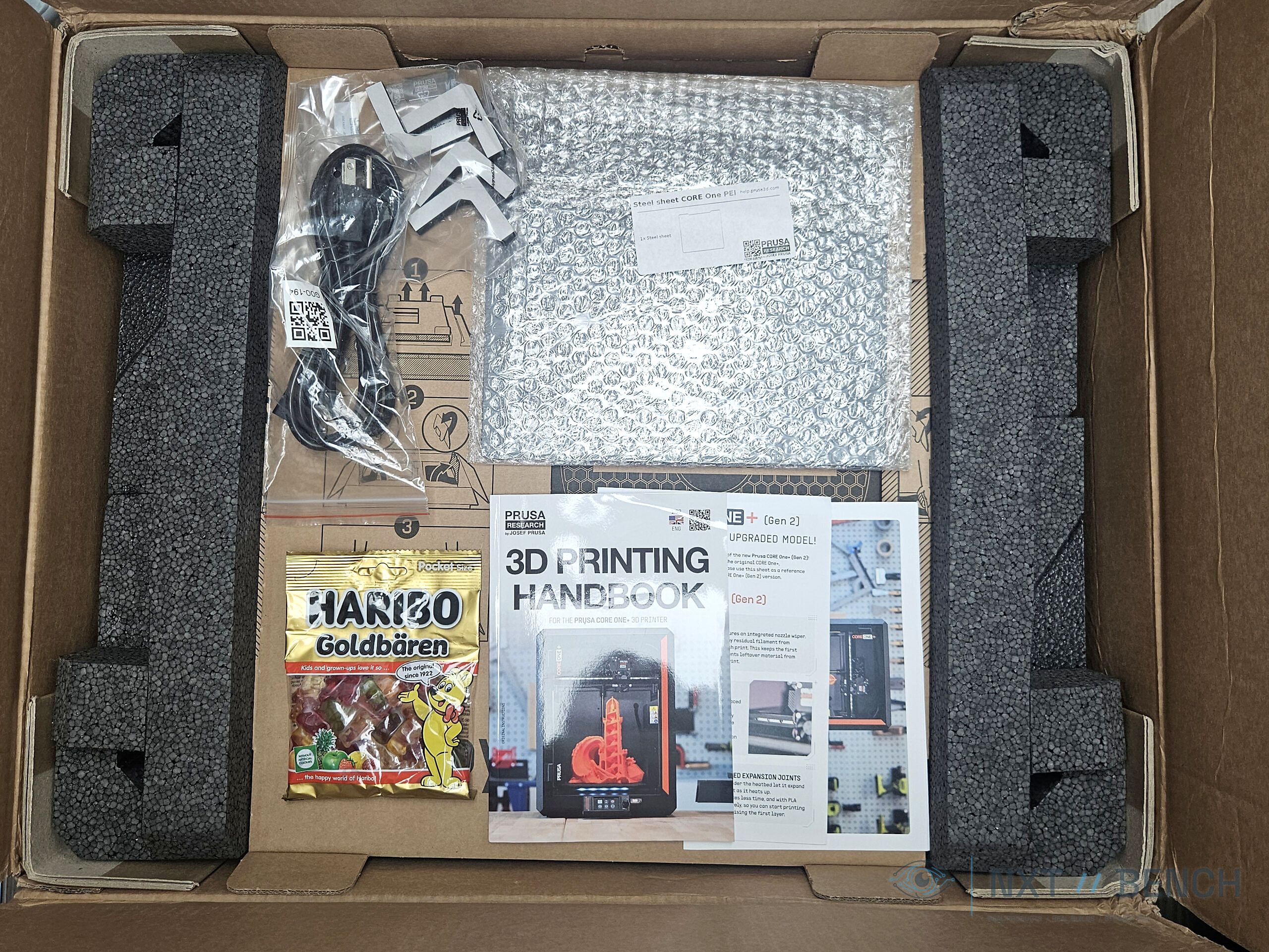 Open Prusa CORE One+ packaging with the 3D Printing Handbook and gummy bears.